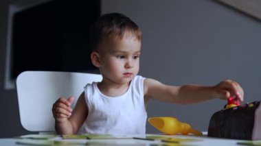 Beyaz tişört giyen bebek oyuna odaklanmış. Ciddi çocuk masasında oturmuş oyuncaklarını oynatıyor..
