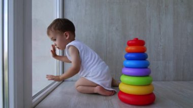 Beyaz elbiseli sevimli küçük çocuk pencerede oturuyor. Küçük oğlan dikkatlice aşağıya bakıyor. Oyuncak piramidi yanında duruyor.