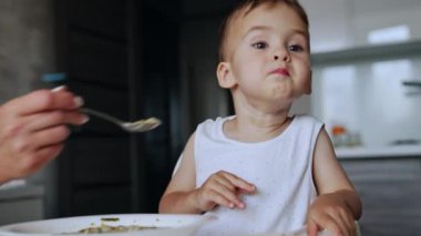 Sevimli erkek bebek yüksek sandalyede oturup geleceğe bakıyor. Annesi ona kaşık veriyor ama çocuk yemek yemeyi reddediyor. Kapatın. Bulanık arkaplan.