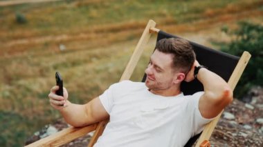 Rahatlamış bir adam katlanan sandalyeye uzanıyor. Telefondaki adam bir elini başının arkasında tutarak görüntülü konuşma yapıyor. Arkaplan bulanık. Üst görünüm.