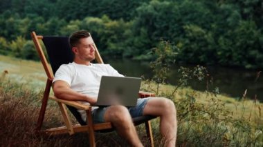 Düşünceli, beyaz bir adam elinde dizüstü bilgisayarla oturup hayal kurarak gülümsüyor. Rahat bir ortamda uzaktan kumandalı çalışan serbest çalışan bir işçi..