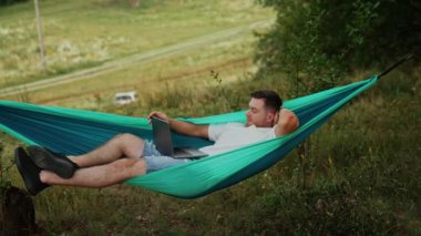 Kafkasyalı adam hamakta yatıyor. Rahatlamış serbest çalışan, ofisten uzakta dizüstü bilgisayarda çalışıyor..