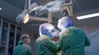 Üç doktor bir cerrahi ekipte çalışıyor. Koruyucu kask takan profesyoneller ameliyat odasındaki lambaların altında yan yana duruyorlar..