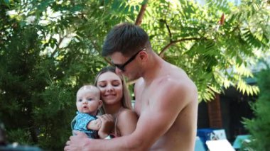 Üç kişilik aile sıcak bir tatile çıktı. Gözlüklü üstsüz bir adam karısını kucağında bir çocukla kucaklıyor..