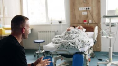 Hastane koğuşunda oturan bir adamın arka görüntüsü. Koca, hamile karısının yattığı yatağa bakar. Bulanık arkaplan.