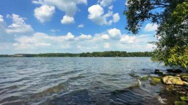 Küçük dalgalar nehir kıyısındaki kayalara doğru yuvarlanıyor. Mavi gökyüzünün altında, yaz günü bulutlu güzel bir su manzarası..