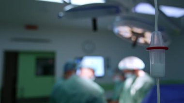 Tezgahın içine beyaz sıvı ilaç damlıyor. Ekipman ameliyathanede. Doktorun ekibi arka planda çalışıyor..