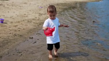 Tatlı çocuk kırmızı sulama kutusuyla su alıyor. Küçük bebek dikkatle huniyi sahildeki kovasına taşıyor..