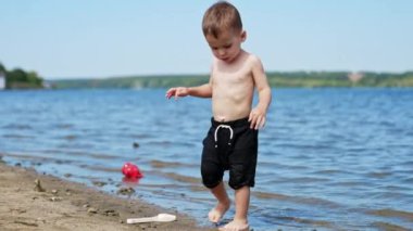 Siyah şortlu şirin çocuk sahilde kumu ve suyu dikkatle takip ediyor. Beyaz çocuk yazın nehir kenarında dinleniyor..