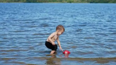 Gülümseyen mutlu çocuk nehirde ayak bileklerine kadar duruyor. Oyuncu çocuk su bidonunu suya atıp yüzüşünü seyrediyor..