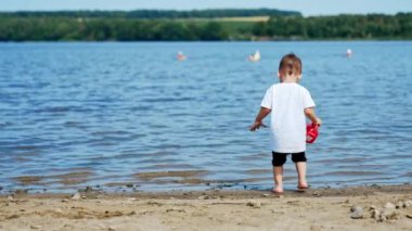 Beyaz tişörtlü sevimli çocuk nehre geliyor. Bebek su bidonunu suyla dolduruyor..