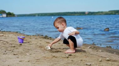 Beyaz tişörtlü beyaz çocuk kumsalda kum kazıyor. Küçük çocuk kıyıda kürekle çalışıyor..