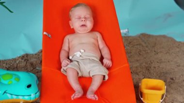 Turuncu sandalyede uzanan gevşemiş bebek. Çocuğun etrafında kum ve yüzme çemberi.