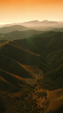 Parlak güneş ışınlarında muazzam kayalık tepeler. Turuncu gökyüzünün arka planında okyanus yakınlarındaki dağlar. Havadan bakış açısı. Dikey video