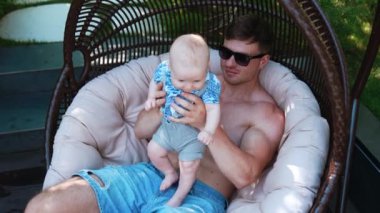 Baba ve küçük oğlu yumurta koltuğunda oturuyorlar. Sevgi dolu bir baba bebeğini sırtından öper. Üst görünüm.