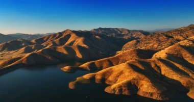 Çıplak kahverengi dağların yamaçları göle doğru iniyor. Batan güneş pürüzsüz kayaları aydınlatıyor. Kaliforniya çöl manzarası.