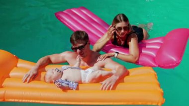 Kafkasyalı ebeveynler bebekleriyle havuzda vakit geçiriyorlar. Bebek yatağında uzanmış, el ve ayak sallıyor..