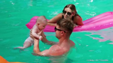 Havuzda küçük bir bebekle yüzmek. Adam oğlunu suyun içinde tutuyor ve annesi yanında yatıyor..