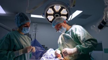 Metal aletlerle ameliyat yapan gözlüklü deneyimli bir erkek cerrah. Hemşire doktor için hazırlanmış bir alet tutuyor. Anestezi uzmanı arka planda duruyor..