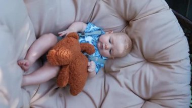 Sevimli beyaz erkek bebek yumurta sandalyesinde rahatça yatıyor. Bebek, elindeki kahverengi oyuncak ayıya bakıyor. - Ne? Dikey ekran.