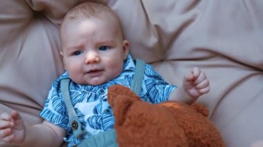 Mavi tişörtlü sevimli beyaz bebek yumuşak arka planda yatıyor. Yumuşak kahverengi ayıcık çocuğun yanında. Kapat..