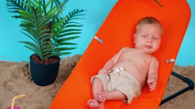 Mavi gözlü sarışın bir bebek sandalyede oturur. Doğaçlama kumsalda şortlu bir bebek dinleniyor..