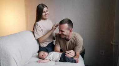 Üç kişilik mutlu bir beyaz aile. Kadın, kocasının saçlarını tarayıp uyuyan bebeğin üzerine oturuyor ve sonra da çocuğun saçını tarıyor..