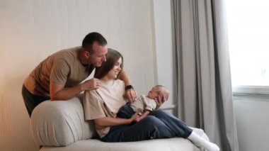 Mutlu aile fotoğrafı. Bej tişörtlü aileler bebeklerinin annesinin ellerinde huzur içinde uyumasına bakıyor..