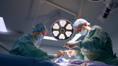 Doktor kesiği dikmeyi bitirir. Kadın hemşire ameliyat ipliğini makasla kesiyor. Düşük açılı perspektif.