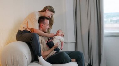 Adam kanepeye oturup yeni doğmuş bir bebeği gözünün önünde tutuyor. Kadın kanepeye oturur, erkeğin arkasında onu öpmek için eğilir..