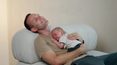 Kanepede elinde yeni doğmuş bir bebekle oturan mutlu beyaz adam. Güzel erkek bebek babasının göğsünde uyuyor..