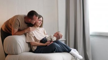 Genç bir kadın, elinde yeni doğmuş bir bebekle kanepede oturuyor. Sevgi dolu koca ve baba kanepenin arkasında onlara yaslandı..