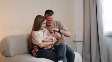 Yeni doğmuş bir bebeği olan beyaz bir aile. Ebeveynler kanepeye oturup annelerinin ellerinde bebeklerine bakarlar. Baba çocuğun elini içine alır..