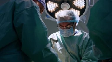 Yetişkin bir erkek cerrah ameliyatta tokmak kullanarak gözlük takıyor. Doktor yuvarlak lambanın altında duran keskiye vurur..