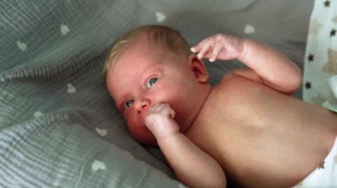 Sevimli beyaz, sarışın, mavi gözlü yeni doğan bebek battaniyelerin içinde yatıyor. Çocuk uyanıkken elini ağzına sokuyor. Kapat..