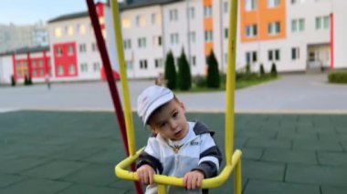 Çocuk parkında sallanan tatlı beyaz bebek. Kameraya bakan sevimli bir çocuk. Arkaplanda bulanık binalar.