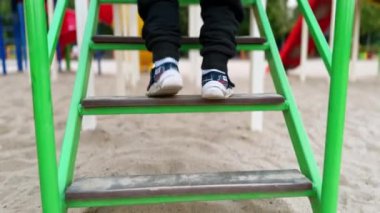 Küçük çocuğun ayakları merdivenlerden yukarı çıkıyor. Çocuk parkta kaydırağın yanında yürüyor. Bulanık arkaplan.