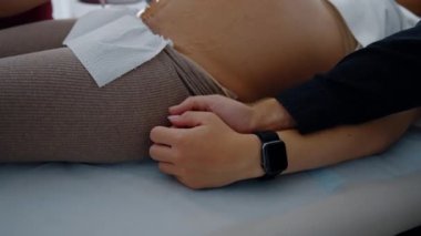 Birbirini kollayan evli çiftlerin elleri. Hamile kadın, ultrason cihazlarıyla muayene edilmiş koltukta yatıyor. Kapat..