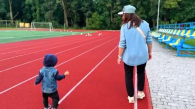 Anne ve oğulun stadyumun yanından geçişlerinin arka görüntüsü. Küçük aktif yürümeye başlayan çocuk pistte koşuyor.