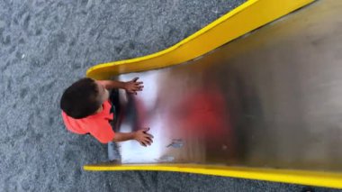 Koyu renk saçlı çocuk metal kaydırağından tırmanmaya çalışıyor. Mutlu çocuk zıplar ve uzaklaşır. Üst Manzara. Dikey ekran.