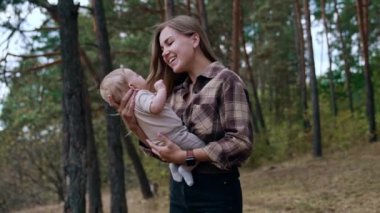 Kareli tişörtlü, bebeğini tutan oldukça uzun saçlı bir kadın. Anne ve bebek doğada vakit geçirirler..