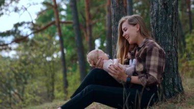 Annem ilk kez bebeğiyle mutlu bir şekilde konuşuyor. Kadın ormanda oturur, ağaca yaslanır ve bebeğine bakar..