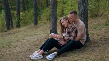 Genç ebeveynler doğada bebekleriyle iyi vakit geçiriyorlar. Ellerinde çocuğu olan bir kadın, ağaca yaslanmış kocasına yaslanır..