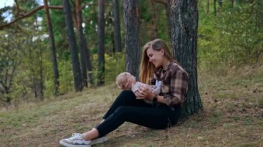 Uzun saçlı mutlu beyaz kadın, kucağında bebeğini tutarak ağaçta oturuyor. Anne konuşuyor ve sevimli küçük oğluna gülümsüyor..