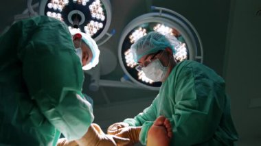 Genç cerrah hastanın ayağını tutuyor ve yaşlı cerrah da aleti uyguluyor. Ameliyat yapan doktorlara düşük açılı bakış açısı.