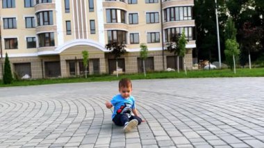 Açık havada oturan beyaz çocuk tişört ve pantolon giyip oturuyordu. Çocuk ayağa kalkar ve mutlu bir şekilde kaçar..