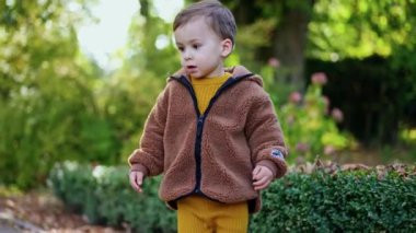 Kahverengi ceket giyen sevimli bebek doğada yürüyor. Çocuk eğildi ve yeşil yapraklara dokundu. Arkaplanda bulanık doğa.