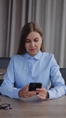 Mavi gömlek giyen uzun saçlı bayan masada oturur. Kadın telefonuna bakar ve not defterine not alır. Dikey video