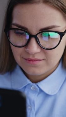 Sarı saçlı, gözlüklü, elinde telefon tutan ve araştıran bir kadın. Telefonuyla çalışan bir bayanın portresi. Kapatın. Yüksek açı. Dikey video