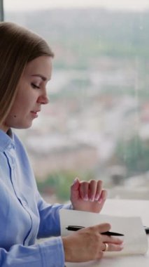 Bayan ofis çalışanı dizüstü bilgisayarla masada oturuyor. Kadın gülümseyen kağıt defterine bakıyor. Arkaplan bulanık. Dikey video